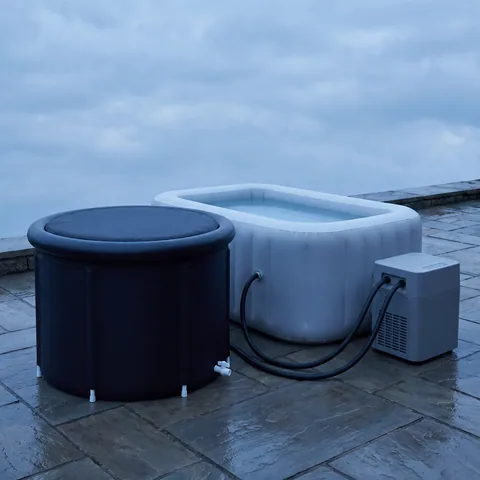 The black round foldable tub next to an unbranded gray chiller connected by two hoses, on a patio, the size difference clearly visible, with no text.
