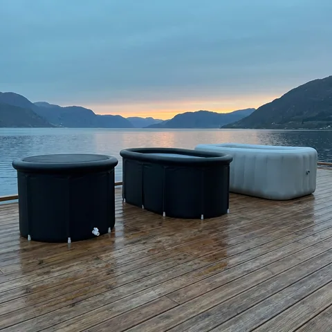 Three kinds of cold plunge tubs side by side on a wooden deck: a black round foldable tub, a longer oval foldable tub and a gray rigid inflatable tub, with no logos