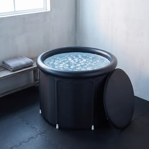 The black round Ice Bath Tub in the corner of a bright home gym set up in a garage, lid off and leaning against it, ice cubes floating on the water, a folded towel on a wooden bench and rubber floor tiles.