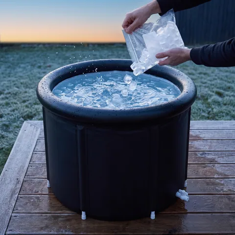 Clear ice cubes poured from a bag into cold water inside the black rim of the tub at dawn, seen from above at an angle.