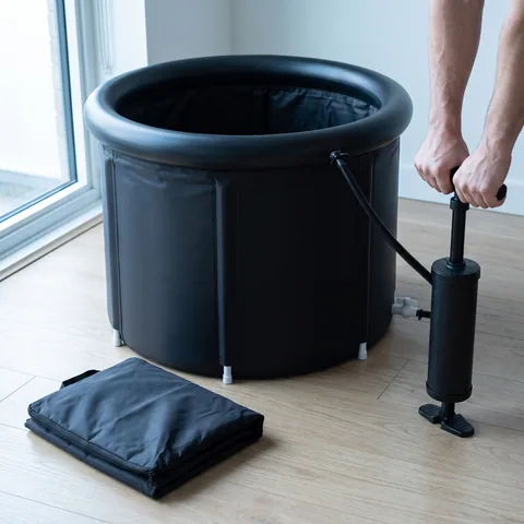 Close-up of the black foldable Ice Bath Tub being set up on a light wooden floor: the hand pump connected to the top ring, the six vertical sleeves of the wall and the folded carry bag beside it.