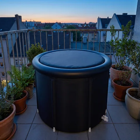 The round black ice bath tub, padded lid in place, on a small city balcony at blue hour, between potted plants, with the top ring and the six vertical seams clearly visible.