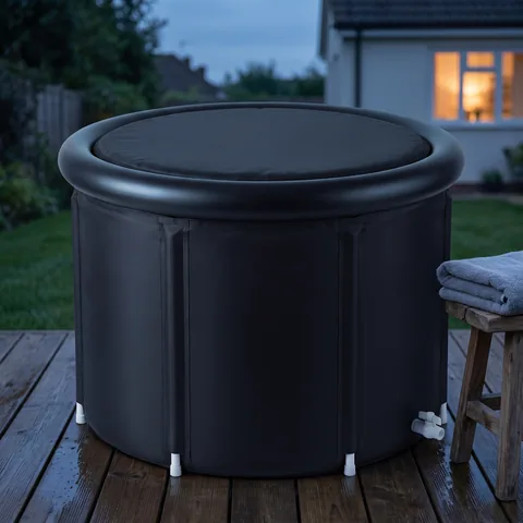 The black round ice bath tub, padded lid closed, on a quiet backyard patio at dusk, with a folded towel on a stool beside it.