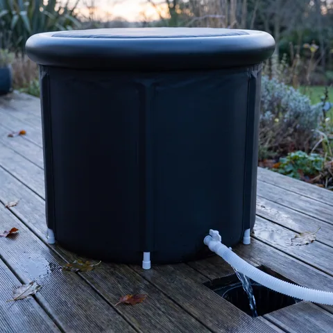 Clear water running through a white hose from the black tub to a backyard drain, over wet deck boards in the morning light