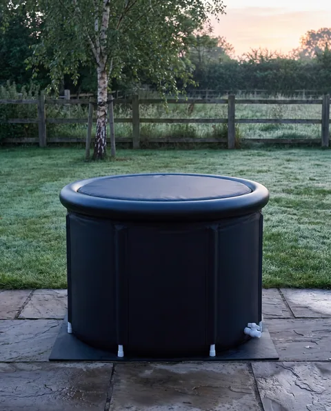 The Ice Bath Tub with its lid closed on a dark mat on a flagstone patio at the edge of a backyard, early in the morning