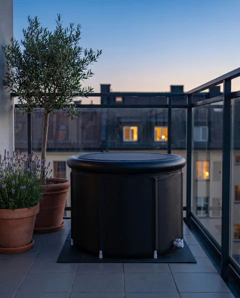 The Ice Bath Tub with its lid on, on a dark mat on a small city balcony with tiles, plants and a glass railing at dusk